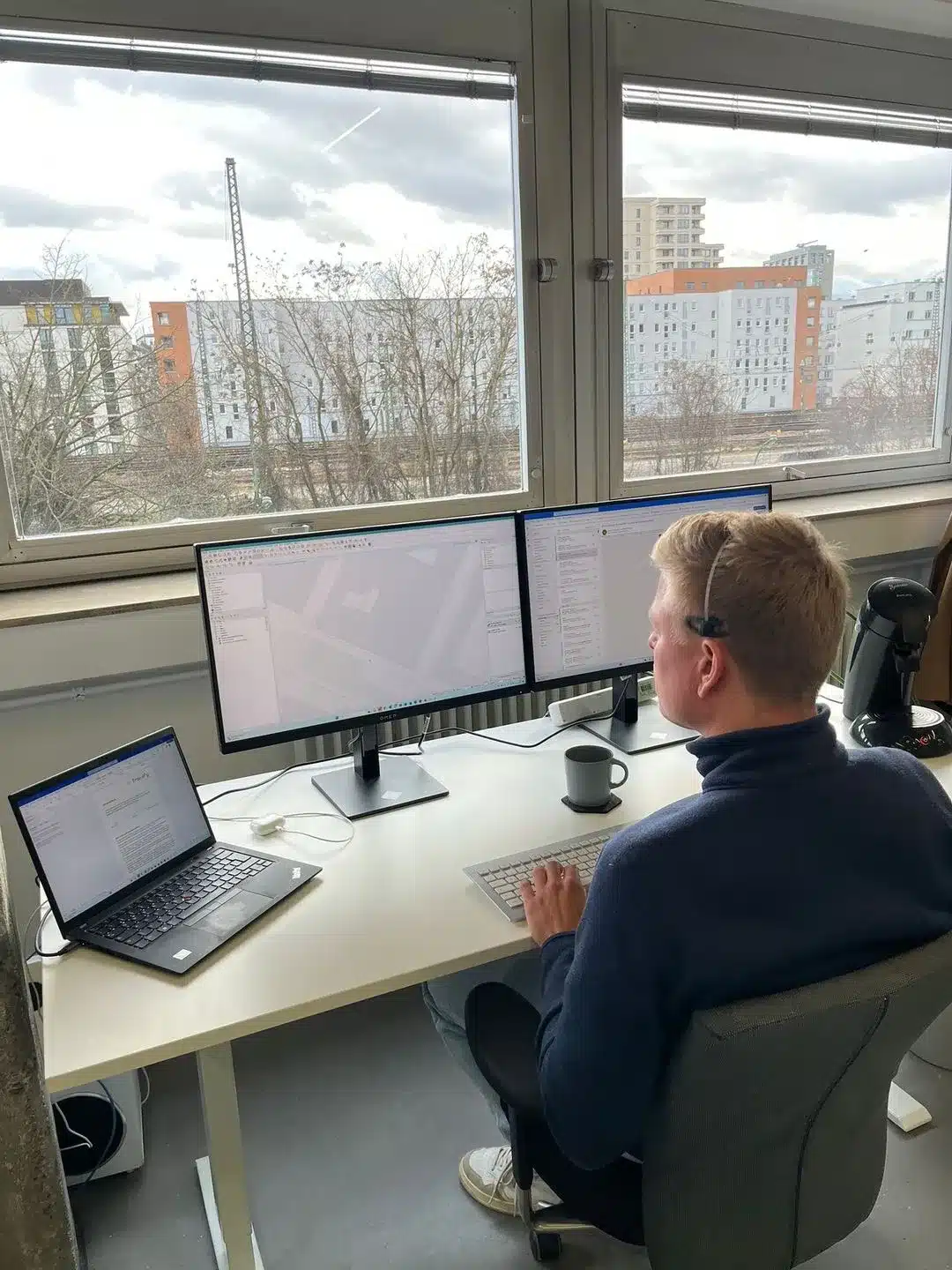 Eine Person sitzt an einem Schreibtisch mit zwei großen Monitoren und einem Laptop und arbeitet effizient an ihrer Online-Leitungsauskunft. Für eine reibungslose Kommunikation trägt sie ein Headset, neben ihr steht eine Kaffeetasse, und durch die großen Fenster sind draußen Gebäude und Bäume zu sehen.