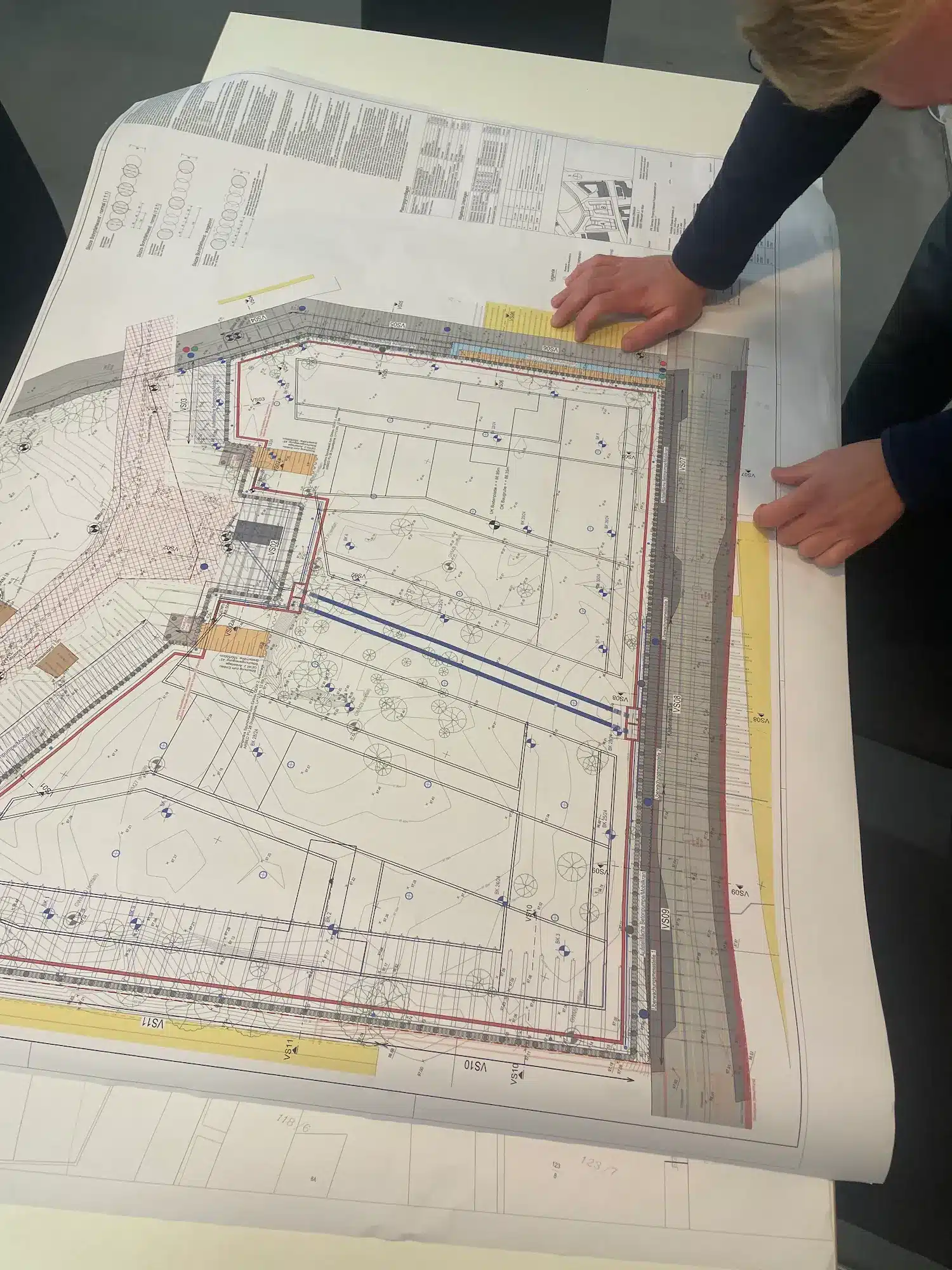 Eine Person sieht sich einen großen, auf einem Tisch ausgebreiteten Architekturentwurf an und prüft detaillierte Baupläne und technische Zeichnungen mit verschiedenen Linien, Symbolen und Anmerkungen – vielleicht, um sich auf die Online-Beantragung einer Leitungsauskunft für ihr bevorstehendes Projekt vorzubereiten.