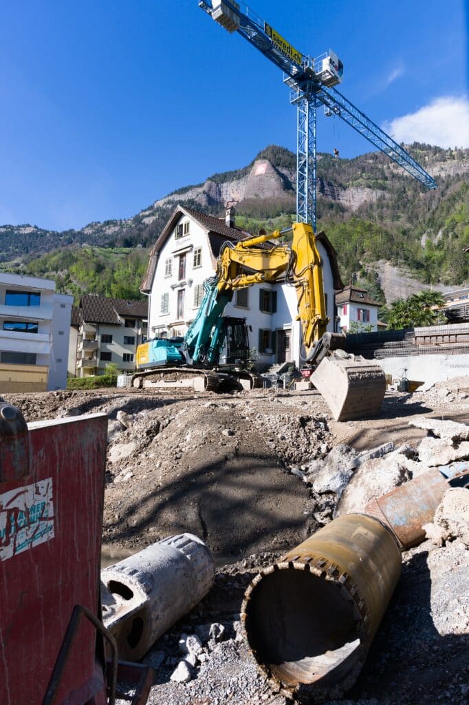 Bagger und Rohre auf einer Baustelle vor einem weißen Haus, im Hintergrund ein hoher Kran und Berge unter blauem Himmel, während Arbeiter die Leitungsauskunft sorgfältig dokumentieren, um einen sicheren und präzisen Baufortschritt zu gewährleisten.