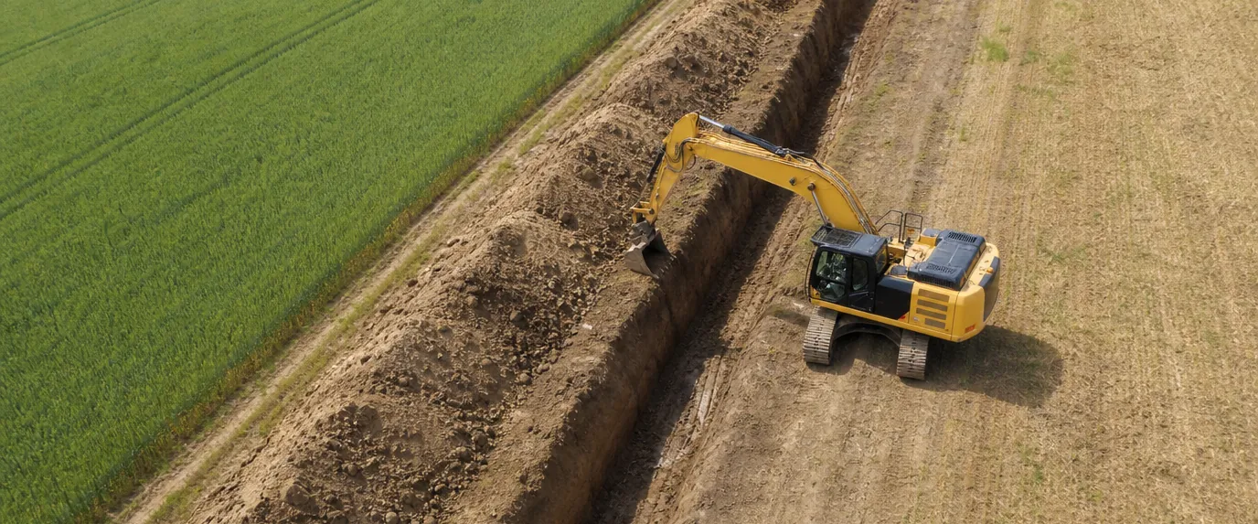 Ein gelber Bagger gräbt einen langen Graben zwischen einem grünen Feld und einem braunen, abgeernteten Feld – aus der Vogelperspektive betrachtet eine typische Szene bei Projekten von Leitungsauskunft Tiefbau.