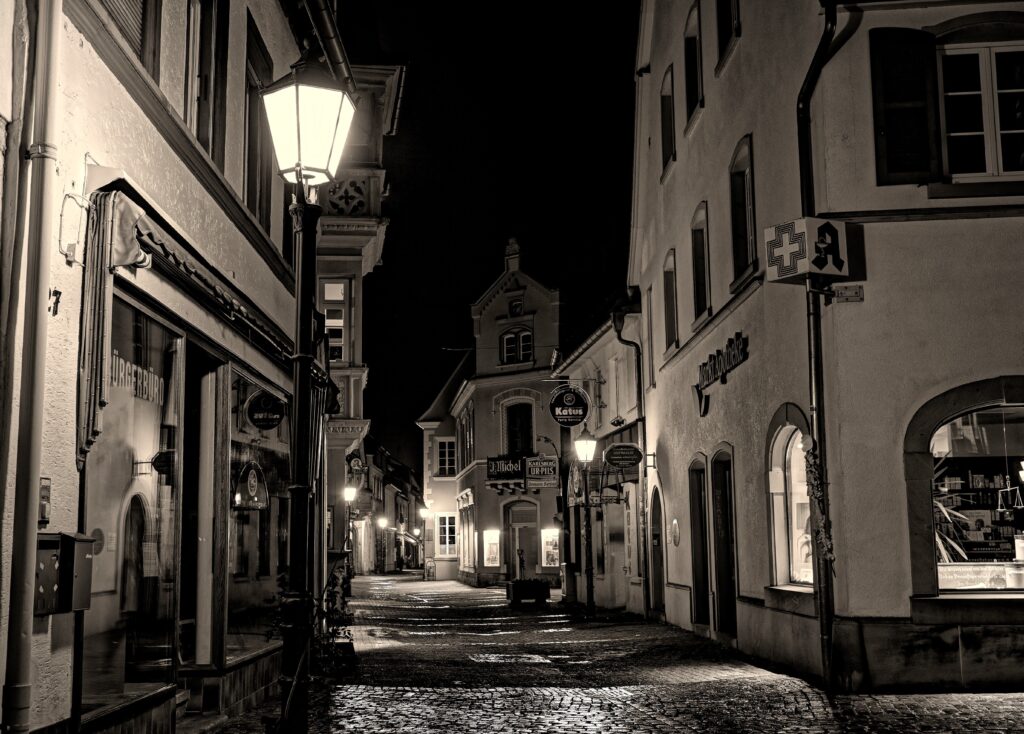 Eine schmale Kopfsteinpflasterstraße in einer europäischen Stadt bei Nacht wird von Straßenlaternen beleuchtet; alte Gebäude, Ladenschilder und das Apothekensymbol sind zu sehen. Das Bild ist in Schwarz-Weiß gehalten.
